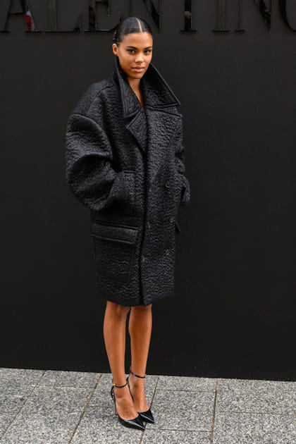 Tina Kunakey en manteau noir à manches bouffantes et escarpins vernis Valentino